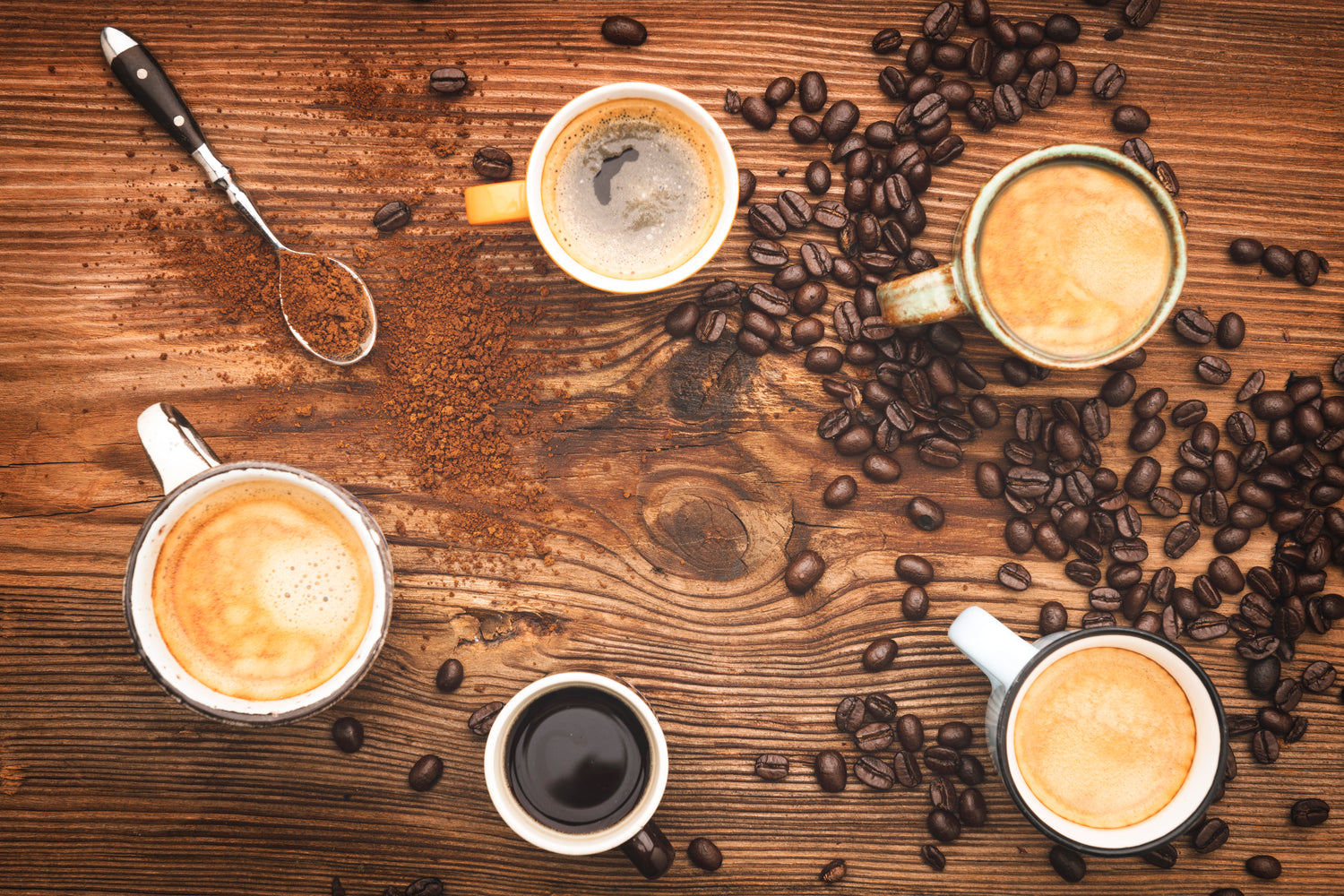 Cups of coffee on a wooden table covered with ground and whole bean coffee.