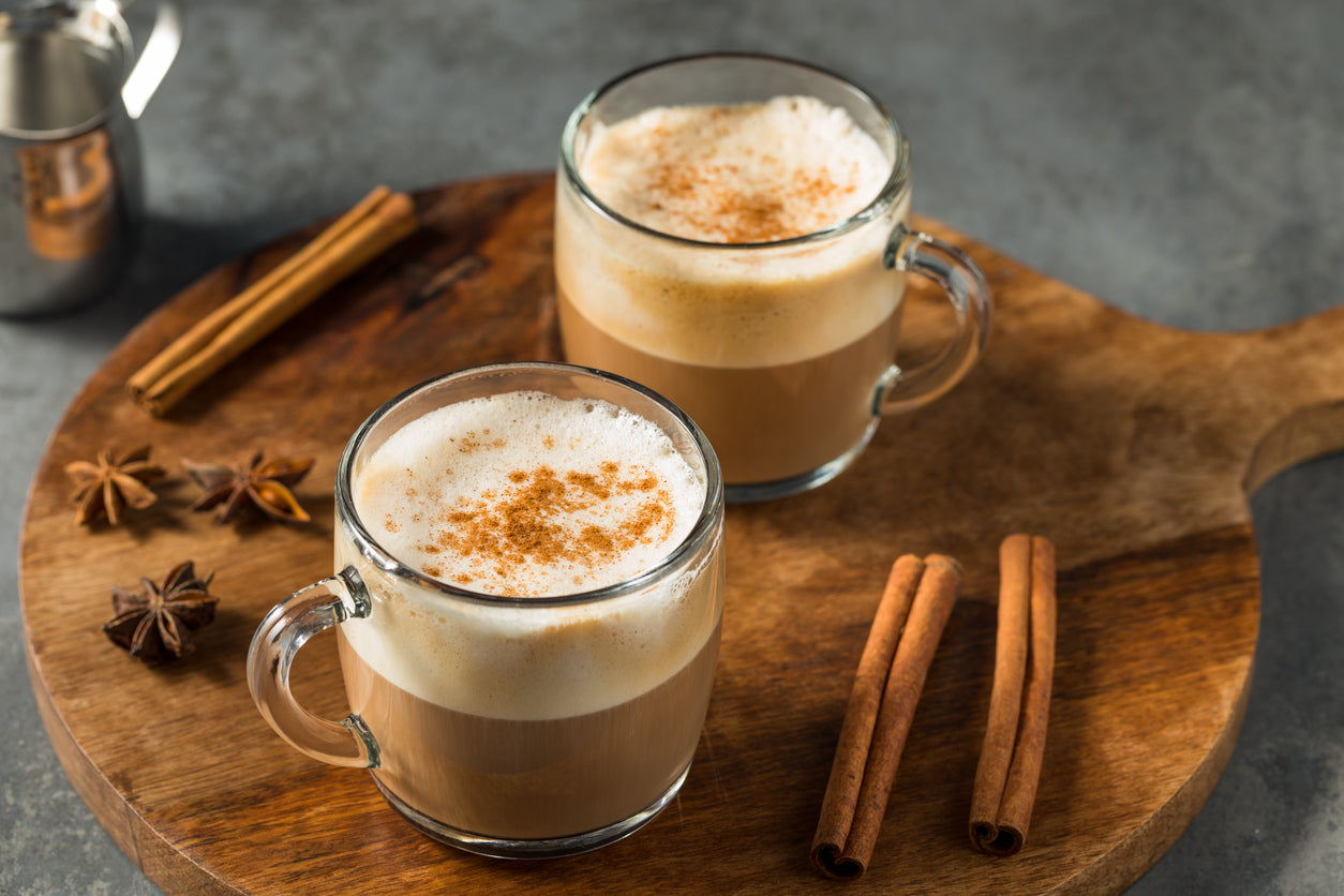 Two cups of coffee in glass cups showing the different layers of the coffee on a wooden tray with cinnamon and star anise.
